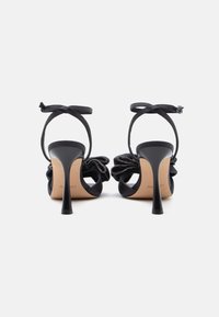 Mustad kõrge kontsaga sandaalid, millel on pahkluuketid ja dekoratiivsed paelad, mis on kaunistatud väikeste helmestega, nähtuna tagantpoolt valgel taustal.