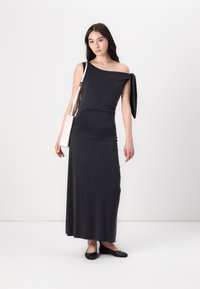 Black, sleeveless maxi dress with an asymmetrical neckline and a tied detail on one shoulder. Model wears black flats and holds a cream bag.