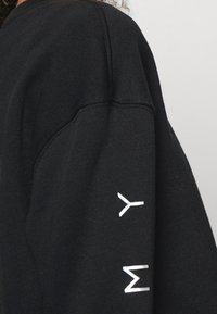 Svart sweatshirt med lös passform, med vita tryckta bokstäver "m y" på ärmslutet. Materialet verkar mjukt med en texturerad yta.