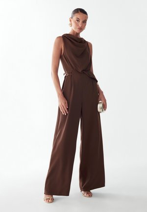 Kvinde iført en ærmeløs brun jumpsuit med vide ben og draperet halsudskæring, der holder en lille metallic clutch og bærer guldringe øreringe og hæle.