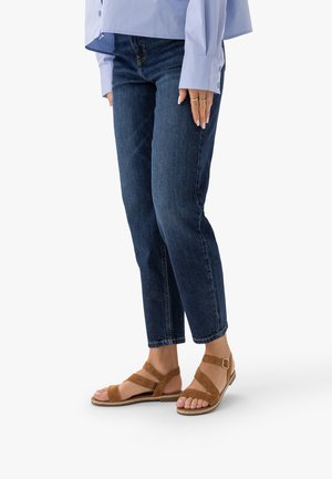 Kobieta w ciemnoniebieskich dżinsach i beżowych zamszowych płaskich sandałach z paskami wokół kostek, stojąca na białym tle.