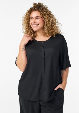 Šypsanti moteris su garbanotais plaukais dėvi juodą, susegamą palaidinę ir juodus kelnes, viena ranka liesdama plaukus, o kita kišenėje.