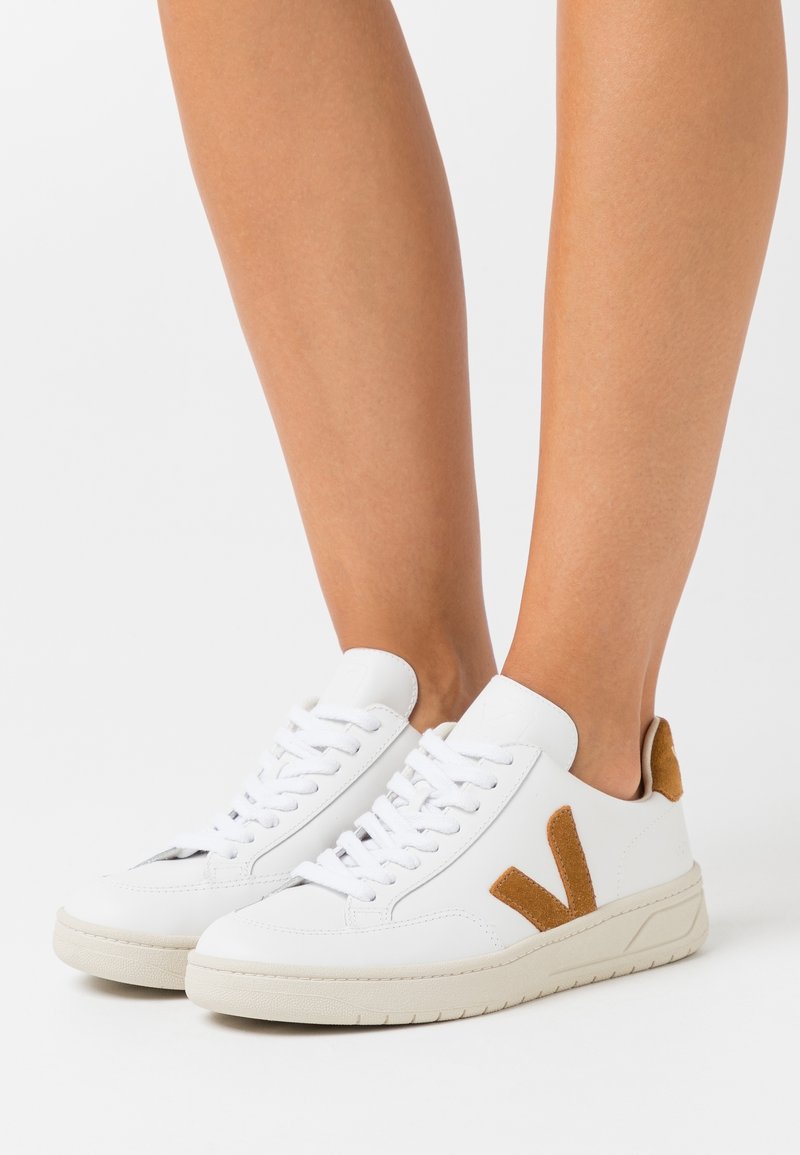 Personne portant des baskets blanches avec des détails en suède beige, dont un logo latéral et une languette au talon, debout sur un fond blanc.