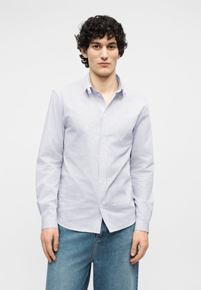 Νέος άνδρας με σγουρά μαλλιά, που φοράει φωτεινό μπλε ριγωτό πουκάμισο με κουμπιά και μπλε τζιν, σε απλό λευκό φόντο.