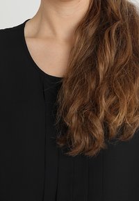 Top nero con scollatura rotonda e dettagli plissettati davanti. Capelli lunghi e ondulati di colore castano visibili, che incorniciano la scollatura.