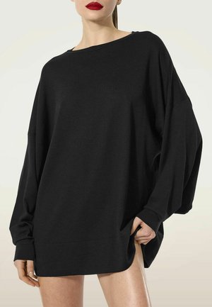Donna con maglione nero oversize a maniche lunghe, rossetto rosso, che tiene l'orlo con una mano su uno sfondo chiaro uniforme.