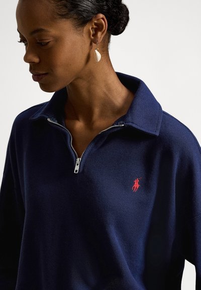 Γυναίκα που φοράει μπλε ναυτικό πουλόβερ με φερμουάρ μέχρι το στήθος και κόκκινο κεντημένο λογότυπο παίκτη πόλο, με κρεμαστό λευκό σκουλαρίκι, κοιτάζοντας προς τα κάτω.