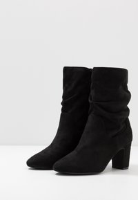 Černé semišové kotníkové boty s volným designem, špičatou špičkou a středním blokovým podpatkem. Mají hladkou texturu a minimální detaily.