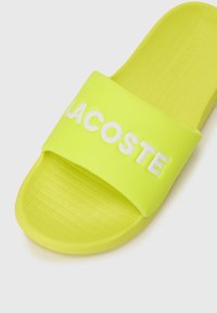 Spilgtā dzeltenā Lacoste slaidu sandale ar teksturētu zoli un platu siksnu, kurā baltiem burtiem ir attēlots zīmola nosaukums.