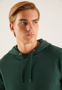Hanorac de culoare verde închis, fabricat dintr-un material moale, având glugă cu șnur și cu gauri metalice. Textură netedă, croială lejeră, design minimalist.