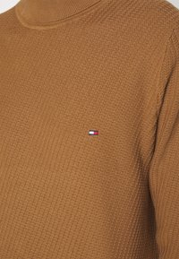 Pull en maille marron texturé avec un petit logo rectangulaire brodé rouge, blanc et bleu sur la poitrine.