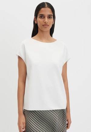 Frau mit langen dunklen Haaren trägt ein schlichtes weißes kurzärmeliges Oberteil und einen schwarz-weiß gemusterten Rock, steht vor einem einfarbigen hellen Hintergrund.