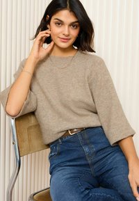 Beige gestrickter Pullover mit Dreiviertelärmeln, kombiniert mit blauen hochgeschnittenen Jeans. Das Modell trägt eine filigrane goldene Kette und ein Armband.
