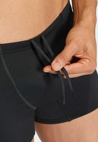 Bañador negro con una textura suave, que presenta un cierre con cordón y costuras en contraste. Acercamiento de una mano ajustando el cordón.