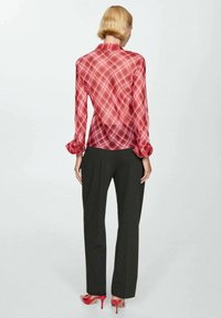 Blusa di raso rossa con motivo a quadretti, dotata di polsini con bottoni e colletto, abbinata a pantaloni grigio scuro e tacchi rossi.