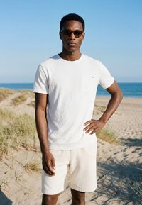 Homme portant des lunettes de soleil, un t-shirt blanc et un short, debout avec une main sur la hanche sur une plage de sable avec de l'herbe et l'océan en arrière-plan.