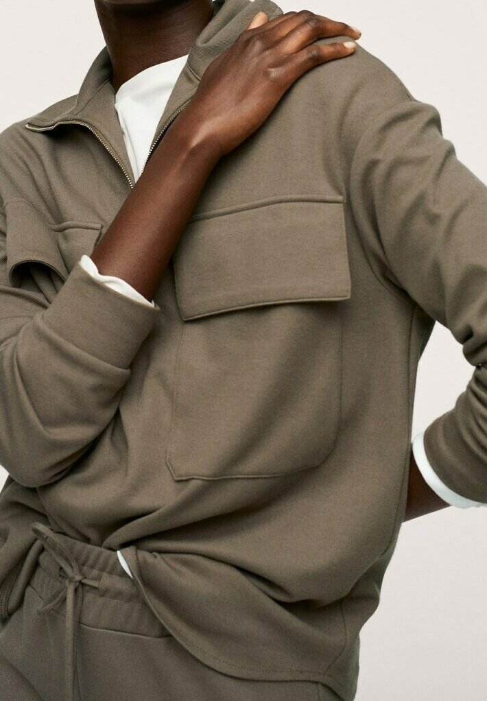 Sudadera marrón clara con cremallera, gran bolsillo frontal, puños acanalados y textura suave. Lleva una camisa blanca debajo.