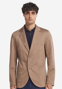 Ljusbrun kavaj i slätt tyg, med slagkrage, två knappar och framfickor. Bärs över en mörk polo t-shirt.