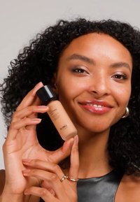 Donna sorridente con capelli ricci tiene una bottiglia di fondotinta liquido KIKO vicino al viso, indossando orecchini a cerchio dorati e una maglietta nera.