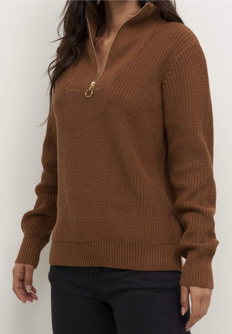 Femme portant un pull en tricot marron avec une fermeture éclair dorée sur un quart, associé à un pantalon noir, debout devant un fond uni.