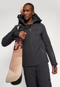 Černá lyžařská bunda s kapucí, zipovými kapsami a logovými detaily, kterou nosí osoba držící hnědou a červenou snowboardovou desku horizontálně.