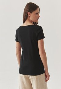 T-shirt noir en coton à manches courtes avec un col légèrement évasé, une coupe ajustée et une texture lisse, porté avec un pantalon en lin clair.
