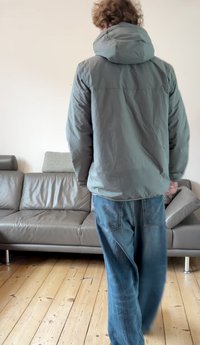 Szara, ocieplana kurtka z kapturem, o gładkiej fakturze i minimalnej ilości szwów, zestawiona z niebieskimi dżinsami. W tle znajduje się skórzana sofa.