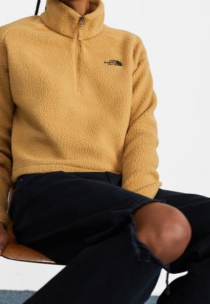 Person iført en beige fleecepullover med kvartlynlås og sorte revnede jeans, siddende på en træstol mod en ensfarvet baggrund.