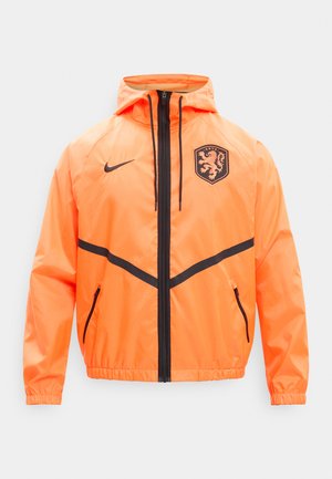 Φωτεινό πορτοκαλί μπουφάν με κουκούλα και μαύρο φερμουάρ, διαγώνιες ρίγες, λογότυπο Nike στο αριστερό στήθος και έμβλημα λιονταριού στο δεξί στήθος.