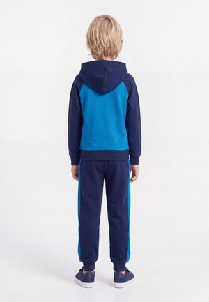 Niño con pelo rubio de espaldas, lleva una sudadera con capucha azul y azul marino, pantalones deportivos a juego y zapatillas azul marino.