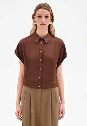 Donna che indossa una camicetta marrone a maniche corte con bottoni e colletto, e pantaloni beige a pieghe vita alta, in piedi davanti a uno sfondo semplice.