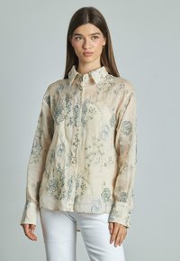 Camicia di lino beige con ricami floreali blu, design a bottoni, maniche lunghe e polsini, che mette in mostra un tessuto leggero e testurizzato.