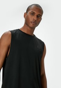 Svart ärmlös träningslinne gjort av lättviktsmaterial, med rund hals och sidoflikar, samt en subtil logotyp på bröstet.