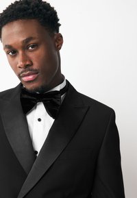 Black tuxedo jacket with satin lapels, white dress shirt, and black velvet bow tie. Smooth fabric, single button closure, and faux pocket detail.