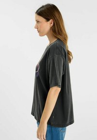 Camiseta gris de gran tamaño con mangas cortas, de textura suave, con un diseño gráfico multicolor en la parte frontal, vista de lado mostrada.