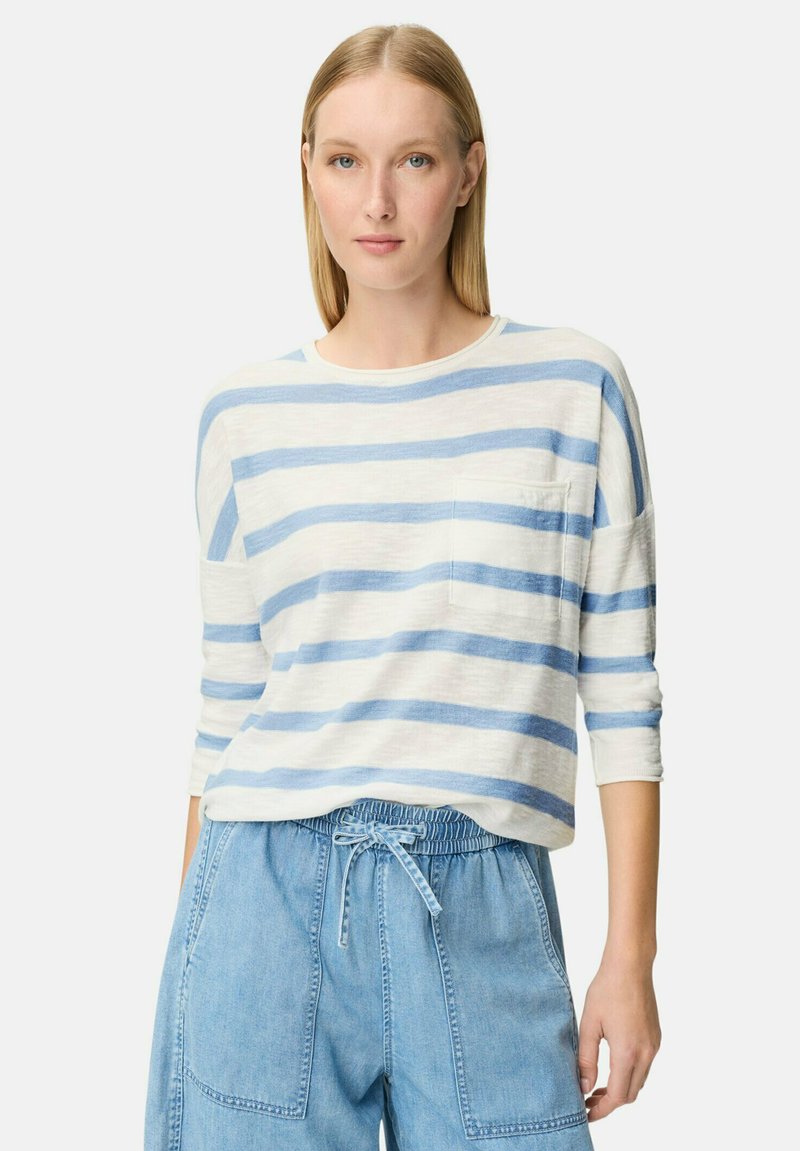 Kvinde med glat blondt hår iført en hvid og lyseblå stribet langærmet skjorte og lyseblå denimbukser med snor i taljen.