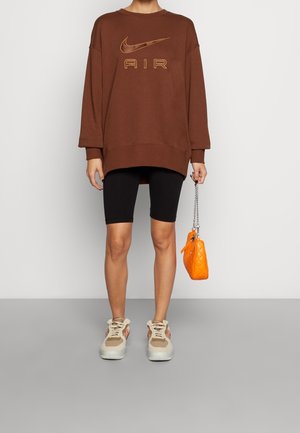 Brązowy oversize'owy sweter z złotym logo "AIR", czarne szorty motocyklowe, beżowe i różowe sneakersy oraz mała pikowana pomarańczowa torebka.