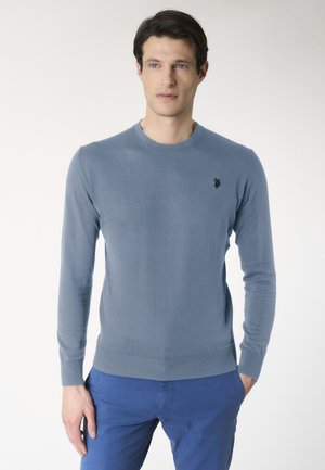 Uomo che indossa un maglione azzurro chiaro a maniche lunghe con un piccolo logo scuro sul petto e pantaloni blu, in piedi davanti a uno sfondo semplice.