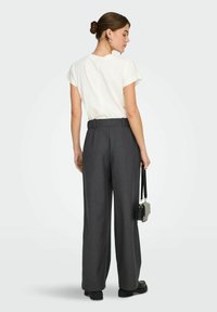 Femme avec les cheveux en chignon portant une chemise blanche à manches courtes, un pantalon gris taille haute à jambes larges, des chaussures noires, tenant un petit sac à main noir.
