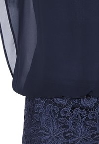 Haut bleu marine en tissu transparent superposé sur une jupe en dentelle ornée de motifs floraux et de broderies texturées. Fini lisse avec un ourlet arrondi.