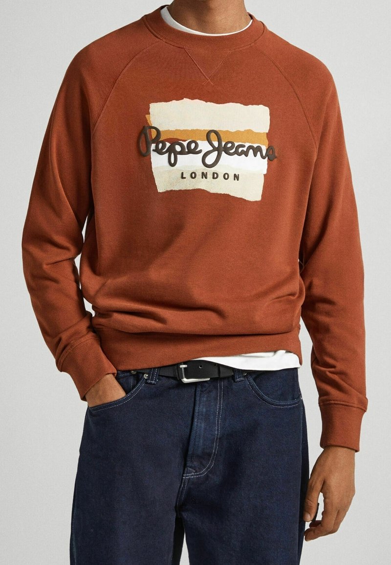Uomo che indossa un maglione color ruggine con il logo "Pepe Jeans London", abbinato a jeans blu scuro e cintura nera.