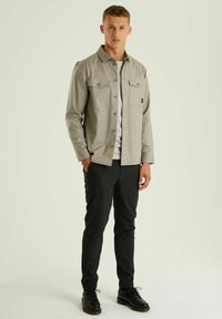 Šviesiai pilkas marškiniai su dviem krūtinės kišenėmis, derinami su tamsiomis liesomis kelnėmis ir juodais batais, stovintys neutralioje aplinkoje.