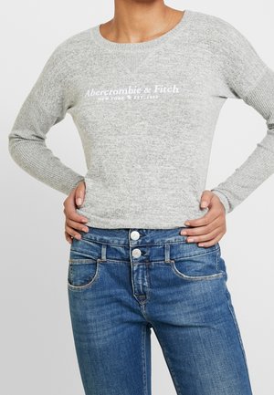 Persoon die een lichtgrijze Abercrombie & Fitch trui draagt en blauwe jeans met dubbele knop, handen rusten op de heupen tegen een effen achtergrond.
