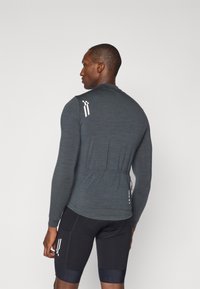 Haut de sport gris à manches longues avec une coupe ajustée, une texture subtile et des accents de marque blancs au dos, présentant un détail de poche arrière.