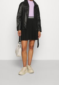 Giacca in pelle nera, top a coste lilla, gonna nera plissettata, sneakers platform beige e piccola borsa beige con tracolla a catena.