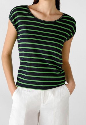 Kobieta w czarnym krótkim topie z zielonymi poziomymi paskami i białych spodniach z wysokim stanem, ręce w kieszeniach.