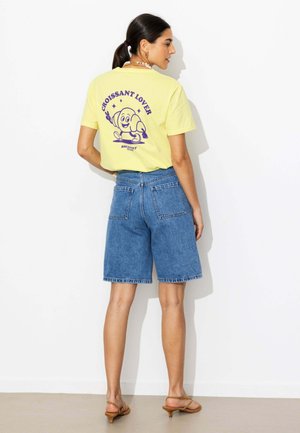 Femme portant un t-shirt jaune avec un croissant violet en dessin animé et le texte « CROISSANT LOVER », associé à un short en jean bleu et des talons beiges.