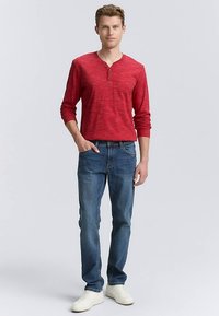 Red long-sleeve henley shirt with subtle texture, paired with blue denim jeans. White sneakers complete the outfit.