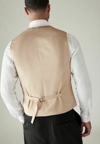 Gilet en satin beige avec une texture lisse, une sangle arrière ajustable et une coupe ajustée, assorti à une chemise blanche à manches longues et un pantalon noir.
