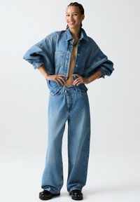 Denimjack tijdens met grote zakken en een ontspannen pasvorm, gecombineerd met brede denim jeans, beide in een lichtblauwe wassing, gedragen met zwarte platform schoenen.